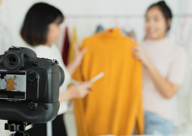Two women hold up a sweater in front of a camera.