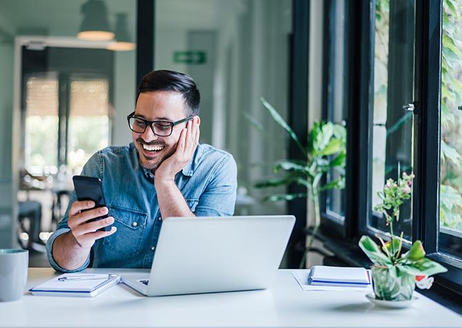 Small Business owners looks at mobile phone smiling while sitting in front of a computer at a desk.